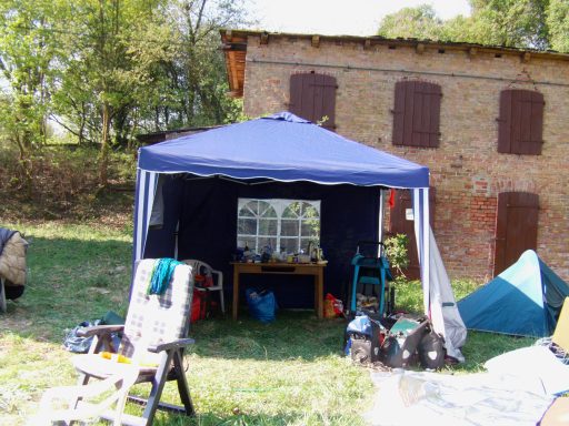 Blaues Zelt mit Tisch und Stühlen auf einer Wiese neben einem alten Gebäude.