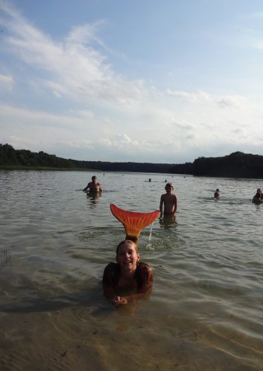 Person im Wasser mit einer orangefarbenen Meerjungfrauenflosse, umgeben von weiteren Schwimmern.