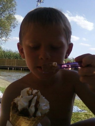 Junge isst ein Eisbecher mit Sahne am Wasser in einer sonnigen Umgebung.